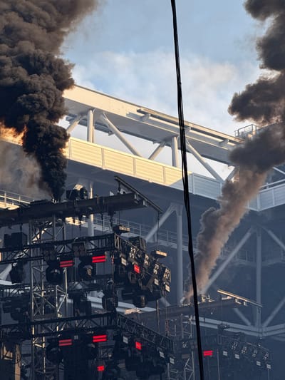 The image captures a dramatic scene, likely an outdoor event or performance in Dusseldorf, Germany. Dark, thick smoke billows into the sky, obscuring the blue sky and clouds with a layer of grey. The smoke appears to originate from the lower left of the frame, with a dense plume extending upwards and to the right, suggesting a significant event, possibly a fire or pyrotechnics.

Dominating the background is a large, industrial-looking structure, likely a stadium or arena, with a complex network of metal scaffolding and lighting rigs. Numerous stage lights are visible, many with red illuminated elements, hinting at a performance setting. The structure itself is a mix of white and grey metal beams and panels. The overall impression is one of intensity and perhaps even chaos, given the presence of the smoke.

The lighting suggests it is daytime, with sunlight illuminating parts of the structure and casting some shadows. The presence of power lines in the foreground adds a mundane contrast to the more dramatic elements of smoke and stage lighting. There is no visible text in the image. The scene evokes a sense of power, perhaps the aftermath of a pyrotechnic display or an unexpected incident at a large gathering.