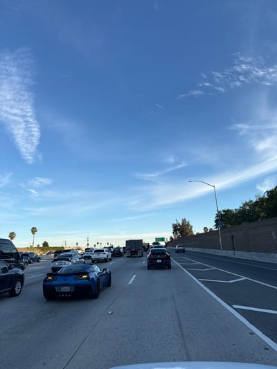 A congested freeway with numerous cars lined up in heavy traffic under a bright blue sky with wispy clouds. A concrete barrier lines one side of the road with trees and foliage visible beyond. A tall light pole stands along the roadside. Palm trees are visible in the distance. The traffic appears to be moving slowly, if at all.