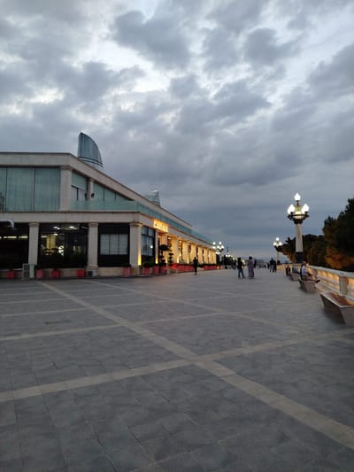 This is a vertical, medium-shot, eye-level photograph of the Baku Boulevard in Sabail Raion, Azerbaijan, taken at dusk. The sky is heavily clouded, with dark gray and lighter gray formations dominating the upper portion of the frame. In the background, a modern building with a distinctive curved glass structure on its roof is visible. The building has large glass windows and is illuminated from within, with a prominent sign reading "MANZARA" in glowing orange letters.

In the foreground, a wide, paved walkway made of dark gray square tiles with lighter gray rectangular inlays stretches into the distance. Several lampposts with ornate, multi-bulb fixtures are spaced along the right side of the walkway. A decorative white balustrade with columns runs parallel to the walkway, separating it from a densely treed area on the far right.

Several individuals are present on the walkway, some walking and others sitting on benches. On the right side, a person is seated on a bench facing away from the camera, looking towards the sea (implied by the view). Further along the walkway, a small group of people are standing and conversing. The overall atmosphere is calm and slightly overcast. The lighting suggests it is either early morning or late evening.