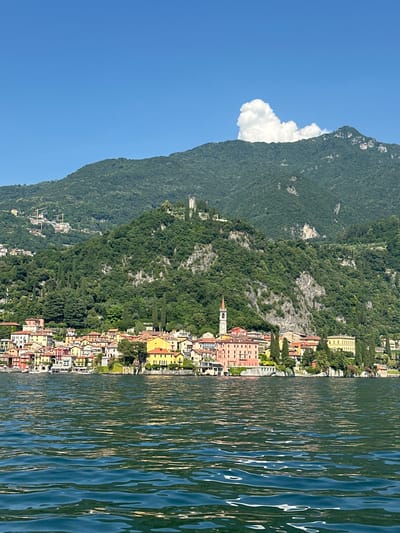 This media file is a vertical, eye-level, wide shot of a picturesque lakeside town nestled at the base of a lush, green mountain. The scene is bathed in bright sunlight, suggesting a clear, warm day. The water in the foreground is a deep blue-green, rippled by gentle waves that catch the sunlight, creating a shimmering effect.
The town itself is a cluster of colorful buildings with red-tiled roofs, stacked closely together along the shoreline. The architecture features a mix of pastel hues, including yellow, pink, and white. A prominent church spire with a clock face rises above the rooftops, indicating a historical and religious presence in the community. Trees, primarily cypress and other evergreens, are interspersed throughout the town and climb the steep slopes of the mountain behind it.
The mountain dominates the background, its slopes covered in dense green foliage. A large, fluffy white cloud is nestled against the mountain, adding a touch of drama to the otherwise clear blue sky. The overall atmosphere is serene and idyllic, evoking a sense of peace and natural beauty. The location appears to be Varenna, Italy, a known tourist destination on Lake Como, which is consistent with the visual cues of the colorful lakeside town and the mountainous backdrop. No people or specific activities are discernible, suggesting a captured moment of landscape and architectural beauty.