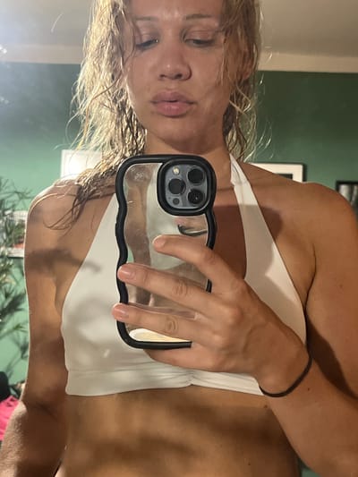 A woman takes a mirror selfie, holding her iPhone. She has wet blonde hair and is wearing a white bikini top. The background shows a wall, possibly in a bathroom, with some greenery visible at the bottom. The time of day and weather are not discernible from the image. The woman appears to be looking at her reflection in the mirror, but her expression is neutral.