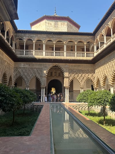 The image depicts the Patio de las Doncellas (Courtyard of the Maidens) in the Real Alcázar of Seville, Spain. The courtyard is an open-air space with a rectangular reflecting pool at its center, surrounded by gardens and arcaded galleries. The architecture is a blend of Mudéjar and Gothic styles, characterized by intricate geometric patterns, horseshoe arches, and detailed tilework.
In the foreground, a long, narrow reflecting pool runs along the central axis of the courtyard. The water is calm, mirroring the surrounding architecture and sky. The pool is bordered by a tiled pathway made of reddish-brown bricks laid in a herringbone pattern, with a thin green tile trim.
Flanking the pool on either side are meticulously manicured gardens with lush green ground cover and several small, leafy trees. These gardens are enclosed by low walls adorned with arched decorative elements, reminiscent of the main building's arches.
The main focus of the image is the stunning architecture of the Real Alcázar. The ground floor features a series of deeply recessed, elaborately carved arches. These arches are supported by slender columns and are adorned with intricate stucco work, showcasing geometric motifs and Arabic calligraphy. Beyond these arches, the building's facade displays further layers of arcades and balconies.
On the upper level, a similar arcade runs along the perimeter, with a white balustrade providing a decorative railing. The wooden ceilings of the arcades are visible, contributing to the historical ambiance.
In the background, a prominent building rises, crowned with a dome and minaret-like structure, its walls decorated with weathered patterns and a reddish roofline. The sky above is a clear, bright blue, with a few birds soaring in the distance.
At the far end of the courtyard, an arched doorway leads into the interior of the Alcázar. A small group of people, appearing to be tourists, are gathered near the entrance, looking into the building or posing for photographs. They are dressed in casual summer attire.
The overall impression is one of serene beauty, rich history, and exquisite craftsmanship. The interplay of light and shadow, the vibrant greens of the gardens, and the intricate details of the architecture create a captivating scene.