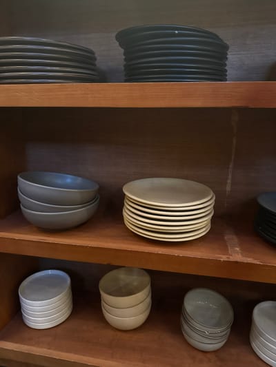 The image shows a wooden shelf containing stacks of plates and bowls. On the left, there are stacks of dark-colored plates. In the center, there is a stack of light-colored plates and a stack of bowls. On the right, there are stacks of bowls and plates in shades of white or light gray. The shelf appears to be part of a built-in structure.
There is no information about the location in the city of San Cristóbal, Mexico, or any information about people, subjects, objects, actions, interactions, and the environment to provide a concise, forensic, matter-of-fact description of the scene.