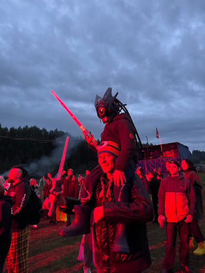 The image captures an outdoor night gathering in a grassy field in Большая Кивара, Russia, illuminated by a dominant red artificial light source. In the foreground, a man with a multicolored beanie (blue, yellow, orange) and a dark jacket carries another individual on his shoulders. The person being carried wears a dark hooded jacket and a prominent black, futuristic helmet with a single glowing red eye and various external components. This individual holds a long, glowing red prop energy sword upright in their right hand and a shorter, similar glowing red prop weapon pointed downwards in their left hand.
To the left of the central figures, a man with dreadlocks, a beard, a dark hoodie bearing the text "Jollity," and plaid pants is partially visible. Further into the mid-ground, numerous individuals are present, some with raised arms, all bathed in the red illumination. A man in a two-tone jacket (dark top, orange/red bottom) and a baseball cap holds a can on the right side of the mid-ground. On the far right, a woman in a dark jacket and bright yellow rubber boots is visible. Smoke, emanating from an unseen source, rises on the left side of the frame, illuminated by the red light. In the background, a makeshift stage or structure, constructed with blue tarps and a tent-like roof, is visible to the right. A flag on a pole stands near this structure. A dense line of trees forms the distant horizon under an overcast, dark sky.