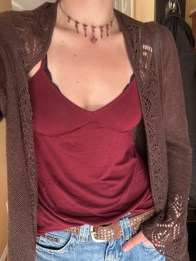 A close-up, angled shot captures the torso of a person, likely reclining or lying down, dressed in a casual outfit indoors. The person's face is not visible, focusing on their clothing and accessories from the chest down to the upper thighs.
The individual wears a dark red or maroon camisole or tank top, featuring a subtle V-neckline. A hint of black lace trim is visible near the bra line. Over this, a dark brown, open-knit cardigan or shrug is draped, featuring intricate lace-like patterns along the sleeves and trim.
Below the red top, light blue denim jeans are visible, along with a prominent brown leather belt adorned with numerous silver studs. A person's hand with unpolished fingernails rests near the belt and jeans.
A silver-toned necklace with multiple red beads or charms hangs around the person's neck, extending onto the upper chest. The person's fair skin has a few small moles visible.
In one of the cropped images, the text "BDG" is visible on the pocket of the jeans. The overall lighting is soft and even, suggesting an indoor setting without specific time-of-day indicators. No specific location cues pointing to Kelowna are visible within the image itself.