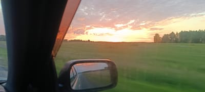 The image depicts a sunset viewed from inside a vehicle. The sun is low on the horizon, casting a warm, orange and pink glow across the sky, which is filled with scattered clouds. A green field stretches out in front of the vehicle, with a line of trees visible in the distance. A side-view mirror reflects the road behind and the sky. The vehicle is on a paved road, and the overall impression is one of travel through a rural landscape. The location is identified as Frolovka, Russia.