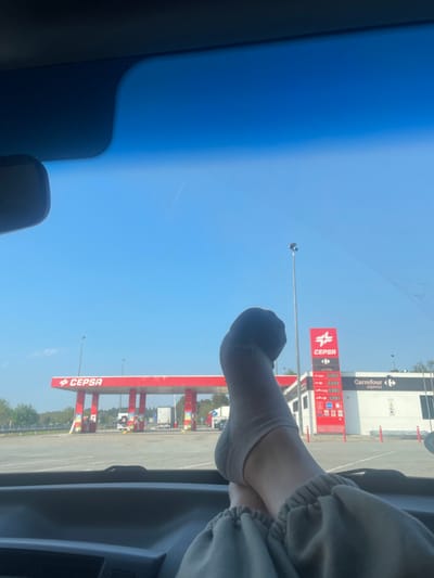 The image depicts a scene from inside a vehicle, looking out through the windshield at a CEPSA gas station and a Carrefour Express convenience store in Garrigàs, Spain. In the immediate foreground, a person's lower legs and feet, clad in light beige ankle socks and grey sweatpants, are visible, propped on the vehicle's dashboard with ankles crossed. Outside the vehicle, a red and white CEPSA gas station canopy, supported by red pillars, covers multiple fuel pumps. To the right of the canopy, a red and white digital display board shows CEPSA branding, fuel prices, and the Carrefour Express logo. Adjacent to this sign, a white building with a "Carrefour express" sign is visible. The station is located on a large paved asphalt lot where several semi-trucks are present in the distance. The environment consists of a clear, cloudless blue sky and a treeline bordering the service area.