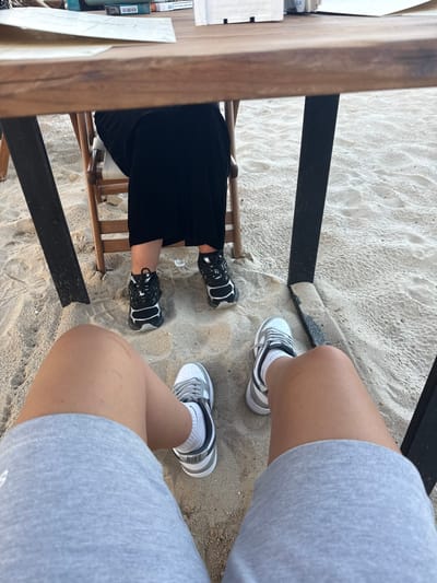 This image depicts two individuals' legs and feet seated on a sandy beach under a wooden table in Kranevo, Bulgaria. The person on the left is wearing black and silver sneakers, and the person on the right is wearing white and gray Nike sneakers with gray shorts. The sand is light in color and textured. A wooden chair is visible between the individuals, and part of a black table leg is in the foreground. In the background, on the table, are what appear to be packs of cigarettes or similar items. The lighting suggests it is daytime.