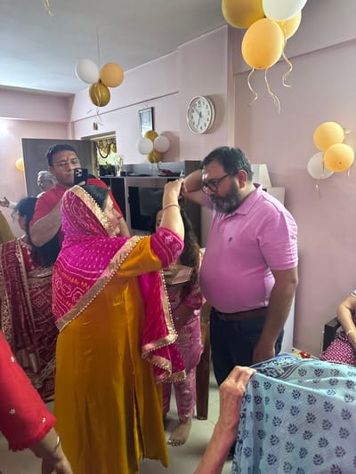 The image depicts an indoor scene in Bidhannagar, India, characterized by a gathering of individuals in a residential setting. A woman, centrally positioned and wearing a yellow kurta with a pink and gold bandhani-style dupatta, has her right arm raised, hand positioned near the forehead of a man standing to her right. The man wears a light pink polo shirt and dark trousers, has a beard and glasses, and his head is slightly bowed with his right hand near his own forehead. Behind the woman, a man in a red t-shirt and glasses holds a black smartphone horizontally, appearing to record or photograph the interaction.

The room features light pink walls and is decorated with clusters of white and gold/yellow balloons suspended from the ceiling and affixed to the walls, indicating a celebratory event. A white circular wall clock with black numerals is visible on the wall to the right of the center. Below the clock, a framed picture hangs on the wall. In the background, a decorated doorway or archway reveals a small shrine or display area with a golden trophy. Partially visible individuals include a person in red attire on the far left, another in pink patterned attire behind the central woman, holding a small tray, and an arm with light blue patterned fabric in the lower right foreground. The flooring appears to be light-colored tiles. The scene is brightly lit.
