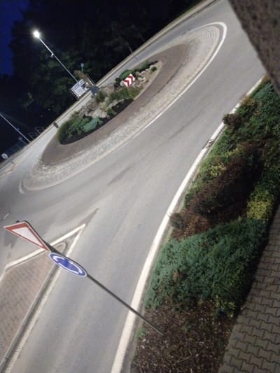 The image shows a street scene at night, likely in Častolovice, Czechia. A roundabout is visible, with a central island featuring rocks and greenery. Road signs, including a give way sign and a roundabout sign, are present. Streetlights illuminate the area, casting shadows on the asphalt. The surrounding environment includes trees, bushes, and paved sidewalks. The perspective is angled from above, giving a clear view of the road layout and surrounding landscape. No people or vehicles are present in the frame.
