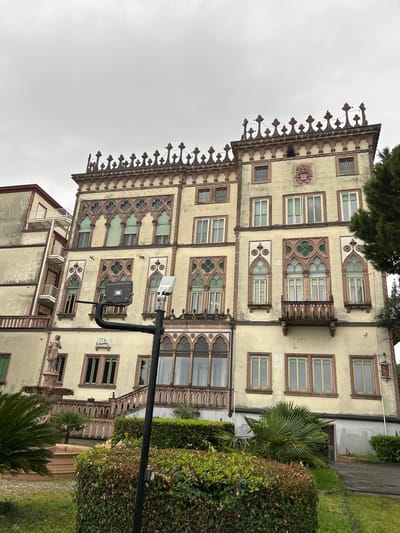 The image shows a multi-story building in Desenzano del Garda, Italy. The building has a yellowish facade with ornate window frames and decorative elements along the roofline. Some windows have green accents, and others are rectangular. A light post is in the foreground, partially obscuring the building. Lush green foliage surrounds the building on the right side. The sky is overcast. Another building is visible in the lower left.