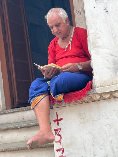 An elderly man with white hair and a fair complexion is sitting on a windowsill in Pushkar, India, reading a book. He is wearing a red t-shirt and blue pants with gold trim. A red cushion with fringe is beneath him. His left leg is crossed over his right, and his bare left foot is visible. The background shows a dark doorway and a white wall with graffiti. The man is holding the book with both hands, his head bowed in concentration. A watch is visible on his left wrist. The scene appears to be outdoors, in a public space.