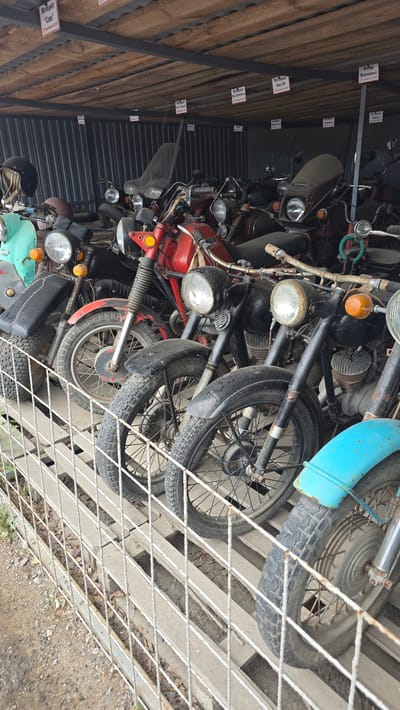 The image depicts a storage area in Bol'shoi Mashlyak, Russia, filled with vintage motorcycles. The motorcycles are lined up in rows on a wooden platform, with a wire fence in the foreground. Several of the motorcycles are red, while others are black, green, and blue. Some of the motorcycles have visible signs with Russian text, indicating their names or origins. The environment appears to be outdoors or in an open-sided structure, with a corrugated metal wall visible in the background. The lighting suggests it is daytime. There are no people visible in the image.