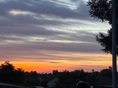 This media file captures a serene sunset scene over a suburban landscape in Sacramento, United States. The sky is the dominant subject, filled with a striking array of colorful clouds. Wispy, elongated clouds streak horizontally across the frame, transitioning from a deep purple and grey at the top to vibrant shades of orange and pink closer to the horizon. The lower clouds appear to be illuminated from below, creating a warm glow that contrasts with the cooler tones above.

Silhouetted against the colorful sky is a line of trees and rooftops, indicating a residential area. The dark foliage of trees frames the bottom of the image, with the peaks of houses visible through the branches. Power lines are also visible, stretching horizontally across the mid-section of the sky, a common urban element. In the distance, a water tower can be seen peeking above the trees, further solidifying the suburban setting.

The time of day is clearly sunset, as indicated by the dramatic coloration of the sky and the low angle of the unseen sun. The weather appears to be clear, with the clouds serving as a canvas for the sunset's spectacle. The overall mood conveyed by the image is one of tranquility and natural beauty. There is no visible text in the media file.