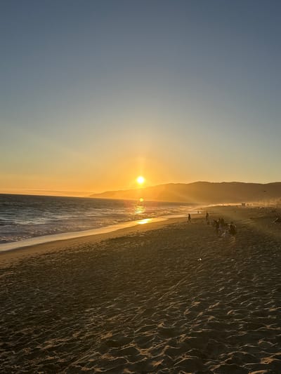 This vertical, eye-level, full shot photograph captures a sunset scene at a beach in Malibu, United States of America. The sun, a bright orb, is setting over the ocean, casting a golden glow across the sky and reflecting on the water. Gentle waves are rolling onto a sandy beach, leaving behind faint footprints. In the distance, rolling hills form a silhouetted backdrop. Several people are scattered across the beach, some walking, others sitting or standing, enjoying the view. The overall atmosphere is serene and picturesque.