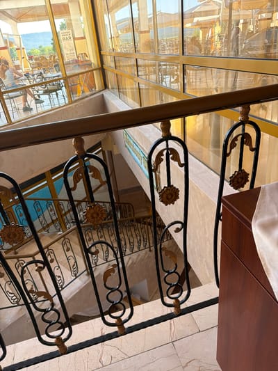 The image shows an indoor view looking down a staircase with ornate black metal railings. The railings feature gold accents and decorative floral motifs. The staircase is lined with light-colored marble-patterned tiles.
Above the staircase, through large windows, an outdoor seating area with tables and chairs is visible, along with umbrellas and distant hills. A sign that reads "SNACK BAR", "WATER", "SMOOTHIES", and "JUICES" is displayed in this area. A woman is seated at a table outdoors.
The overall environment appears to be a hotel or restaurant setting, possibly in the city of Dyuni, Bulgaria, given the context provided. The lighting suggests it is daytime.