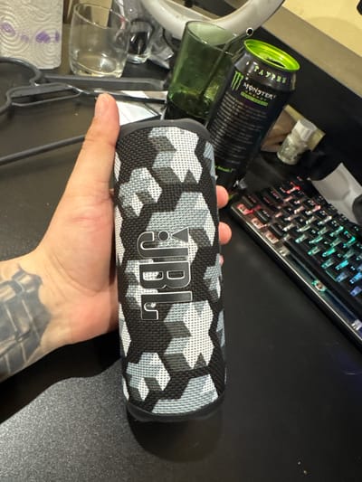 A hand is holding a JBL speaker with a black and white geometric pattern against a cluttered background. The background includes a gaming keyboard, a Monster energy drink can, a glass mug, and miscellaneous items on a dark surface. There's no indication of Kavarna, Bulgaria.