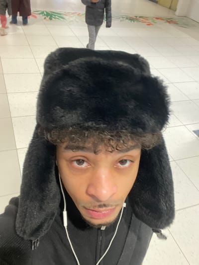 The image depicts an individual situated in an indoor public space within the city of Plaisir, France. The central subject, a person with medium brown skin, dark curly hair visible below a hat, and a slight mustache and soul patch, occupies the lower foreground, looking directly at the camera. They are wearing a large black furry ushanka-style hat, a black ribbed zipped top, and white wired earbuds are visible. The background reveals a light-colored tiled floor with faint decorative green and orange/red leaf-like patterns. Several blurred figures are visible in the mid-ground and background: one figure on the far left wearing a reddish-brown garment and blue footwear, and another figure on the mid-right in a dark puffer jacket and light grey pants, walking away from the camera. The scene suggests a transient public area, such as a shopping center or similar facility.