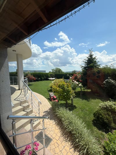The image shows a view of a lush green backyard on a sunny day, located in Aytos, Bulgaria. The backyard features a well-maintained lawn, surrounded by hedges and various plants and trees, including a prominent evergreen tree. In the background, a hammock is visible on the lawn, suggesting a place for relaxation. A red lawnmower is parked on the grass. The scene also includes a part of a building with a curved balcony and a staircase with metal railings. The sky is blue with scattered white clouds.