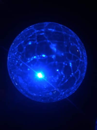 The image shows a close-up, low-light, and slightly blurry view of a transparent, spherical object illuminated from within with bright blue light. The object appears to be made of glass or plastic and has a textured surface, possibly a network of internal veins or cracks, with small, scattered points of light. A brighter, greenish-white light source is visible at the center, with a beam of light extending outwards. The background is completely dark, creating a strong contrast with the illuminated sphere. There is no indication of a specific location in Belashtitsa, Bulgaria, nor are there any people, actions, or interactions depicted. The scene is static and abstract.