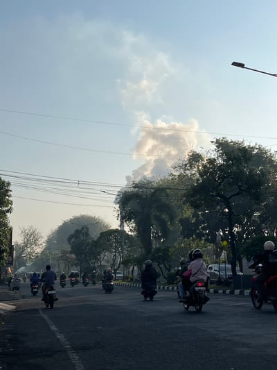 The image captures a street scene in Candi, Indonesia, dominated by a large plume of smoke rising into the clear sky. The smoke appears to be emanating from a distant source, possibly a fire or industrial activity. A road runs through the center of the image, with several motorcycles and their riders navigating the traffic. The motorcycles are traveling in both directions, and some are carrying multiple passengers. The riders are mostly wearing helmets for safety. Trees and foliage line the sides of the road, with some taller palm trees visible. Power lines are strung across the sky, and a street lamp is visible on the right side of the frame. The overall atmosphere suggests a normal day, with the exception of the prominent smoke plume, which could indicate an emergency or ongoing event in the vicinity.