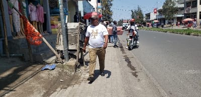 The image shows a man walking down a sidewalk in Rubavu, Rwanda, at coordinates -1.668, 29.268. He is wearing a white t-shirt with a logo, brown pants, and a red baseball cap. He is walking in a relaxed manner and is looking straight ahead. The sidewalk is lined with buildings and trees. There is a motorcycle parked on the street behind him, and several people are visible in the distance. There are two people sitting on a bench on the sidewalk, several people walking down the sidewalk, and a few more people walking in the street in the distance. The scene is sunny, and the sky is clear. There is a street vendor on the sidewalk, and a blue sheet of metal is on the ground in front of him. There is a large red object with a white string hanging from it at the beginning of the sidewalk. The image was likely captured during the day as it is sunny.