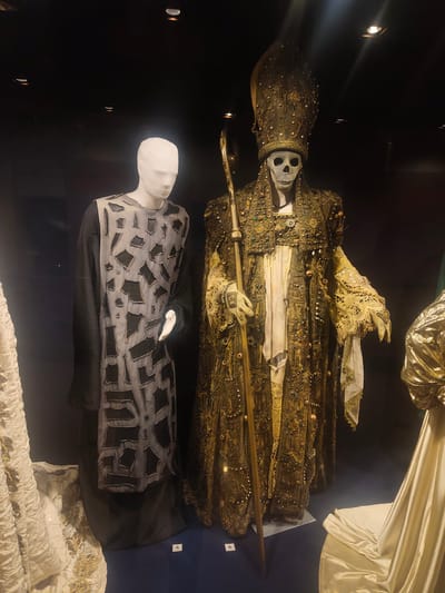 The image depicts two mannequins in a display case, likely in a museum. The mannequin on the left is wearing a dark robe with a lighter, patterned overlay featuring geometric shapes. The mannequin on the right is dressed in an elaborate, dark, and gold ceremonial outfit, including a tall headdress and a skull face. It holds a staff. The background is dark, with spotlights illuminating the display. Numbers "5" and "6" are visible on the display. Based on information provided in the original query, this scene is located in the Museum of Byzantine Culture in Thessaloniki Municipal Unit, Greece.