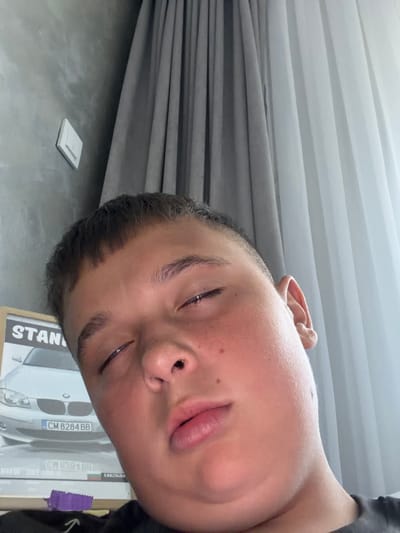 The image is a close-up, high-angle selfie of a young male with fair skin and short, dark hair. He has his eyes partially closed and his mouth slightly open, giving the impression he is tired or drowsy. He is looking downwards at the camera, which is positioned above him. The background consists of a grey, textured wall on the left, with a white light switch visible. To the right, there are light grey, floor-to-ceiling curtains. In the lower left quadrant of the image, a framed poster or advertisement is partially visible, featuring a silver car, likely a BMW, with a Bulgarian license plate reading "CM 8284 BB". The environment appears to be indoors, possibly a bedroom or living space, in a residence located in Zmeitsa, Bulgaria, as indicated by the car's license plate.