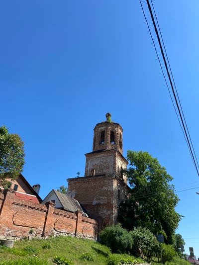 The image displays the dilapidated brick bell tower of the Church of the Dormition of the Theotokos (Uspenskaya Church) in Serpukhov, Russia, against a clear, deep blue sky. The multi-tiered bell tower, constructed primarily of red brick with sections of faded plasterwork, exhibits significant weathering and decay, with green vegetation growing from its upper parts and openings. To the left, a long, aged red brick wall with decorative recessed panels runs along a grassy incline. Behind this wall, the roofs and upper sections of two adjacent buildings, one with a red metal roof and another with a grey sloped roof, are visible. A large, dense green tree partially obstructs the lower right side of the bell tower. In the foreground, more green foliage, including bushes and grassy areas, covers the sloping ground. Three dark utility lines are strung diagonally across the upper right quadrant of the frame. The scene is illuminated by bright daylight, casting subtle shadows. No individuals are present.