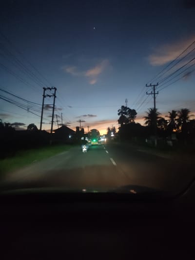 The image captures a twilight scene from a vehicle driving down a paved road in Cotbuket, Indonesia. The sky displays a gradient of deep blue to orange, indicating dusk or dawn. Scattered clouds are illuminated by the setting or rising sun, casting a warm glow. Silhouetted against the sky are utility poles with numerous power lines crisscrossing overhead, typical of a populated or developed area.

On either side of the road, the landscape is lined with dark, indistinct shapes of houses and lush vegetation, including palm trees on the right, suggesting a tropical or subtropical environment. The road itself is illuminated by headlights and streetlights, with two vehicles visible in the distance: a motorcycle with its headlight on, and a truck with its green hazard lights illuminated.

The overall mood is serene and transitional, evoking the quiet moments of travel during the twilight hours. The low light conditions and the presence of vehicles suggest movement and activity, but the scene remains peaceful. There is no visible text in the image.