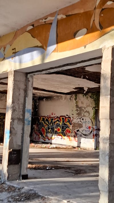 The image, taken from an elevated perspective, depicts the interior of an abandoned structure in Podbadac, Montenegro. Concrete beams and crumbling walls frame a view downward. The walls are covered in graffiti, with tags in black, red, and yellow. There's some scattered debris in the lower area. The scene shows signs of neglect and decay. The image is taken during daytime.