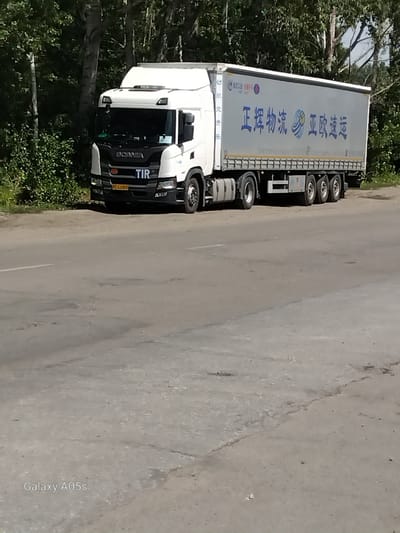 A white semi-trailer truck with "SCANIA" written on the front is parked on the side of a paved road, with trees and foliage visible in the background. The trailer of the truck is white with blue Chinese characters that translate to "Zhenghui Logistics, Asia-Europe Express." A yellow license plate with the characters "C 229RS" is visible on the front of the truck. The road surface is cracked and uneven. The scene appears to be in a rural or suburban area, possibly on the outskirts of Aleysk, Russia. There are no people visible in the image. The time of day appears to be daytime, with sunlight casting shadows. The image is watermarked with "Galaxy A05s".