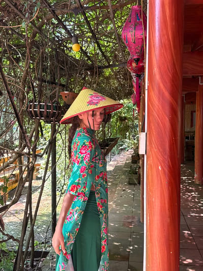 A full-length, portrait-style shot captures a woman standing outdoors in what appears to be a garden or courtyard setting in Thông Nguyên Commune, Vietnam. She is wearing a traditional Vietnamese conical hat (nón lá) with yellow stripes and pink floral patterns. Her attire consists of a long-sleeved top featuring a vibrant green background with bold red and pink floral designs, worn over a plain green dress or skirt. Her long dark hair is styled in a braid that falls over her shoulder, and her facial expression is neutral, with her eyes partially shadowed by the hat.

The background is densely packed with interwoven bare tree branches and green foliage, creating a natural canopy. Among the branches, several decorative lanterns are visible, including a prominent pink one adorned with a bird and bamboo motif, and a red lantern. A single yellow light bulb and a dark, cylindrical hanging object are also interspersed within the foliage.

To the left and top of the frame, a large, reddish-brown wooden beam or pillar from a traditional architectural structure is prominent. The ground beneath the foliage exhibits dappled sunlight, indicating a bright, sunny daytime. The overall scene evokes a serene and culturally rich atmosphere. No visible text is present in the image.