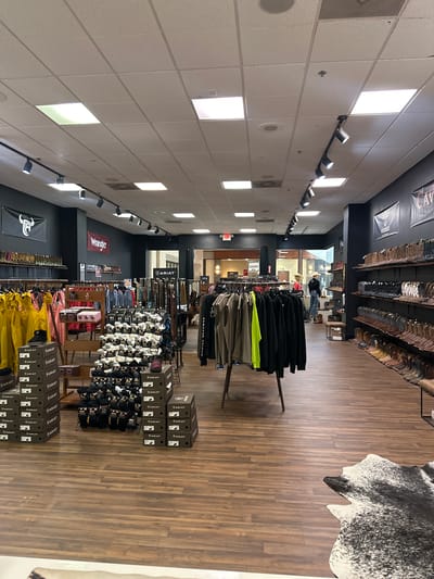 The image shows the interior of a retail store, likely a western wear or boot store, located in Greenville, United States of America. The store is well-lit with overhead fluorescent lighting and track lighting directed at displays. The flooring is made of light brown wood planks.
Various clothing items and accessories are displayed throughout the store. There are racks of pants and jackets, including black jackets and brightly colored pants. Shelves are filled with boxes of socks, many labeled with the brand "ARIAT". The store also features displays of boots on shelves along the walls and on tables. Signs for brands like "Wrangler" and "ARIAT" are visible. A "CLEARANCE" sign is also present.
In the background, a few people are visible, including a person wearing a cowboy hat and another person looking at merchandise. The overall atmosphere appears to be that of a typical retail environment focused on selling apparel and footwear. The time of day is not specified, and the weather is not discernible from the indoor setting. The arrangement of merchandise suggests an organized and well-stocked store.