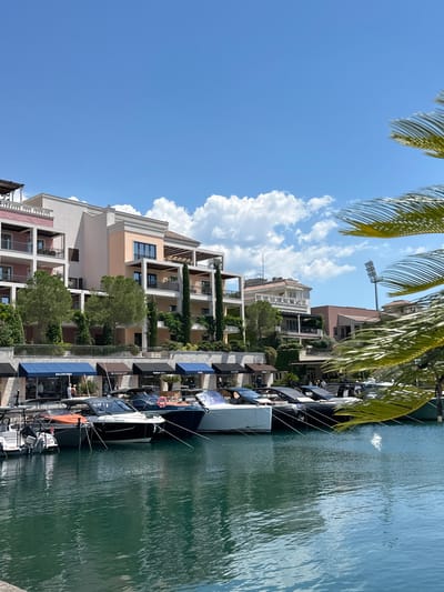 This image depicts a scenic view of a marina in Donja Lastva, Montenegro, under a clear blue sky with scattered white clouds. The foreground is dominated by the calm, dark green water of the marina, with several modern motor yachts docked along the edge. These yachts vary in size and color, with prominent brands like Balenciaga, Givenchy, and Fendi visible on the awnings of the shops lining the waterfront. Behind the yachts, a row of multi-story buildings with balconies and terraces overlooks the marina. These buildings are designed in a contemporary architectural style, featuring a mix of pastel colors and white accents, with lush greenery and tall, slender trees planted around them. In the background, a tall stadium light fixture stands out against the sky, suggesting a nearby sports facility. The overall scene conveys a sense of luxury and leisure in a waterfront setting.