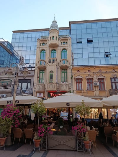 The image depicts a street scene in Niš, Serbia, with a prominent focus on the facade of a building that exhibits a juxtaposition of old and new architecture. A traditional, ornate building with a cream-colored facade and green-trimmed windows stands beside a modern building clad in reflective blue glass. In the foreground, several large white umbrellas, branded with the "PEPSI" logo, are set up, indicating an outdoor seating area for a cafe or restaurant. Wicker chairs and tables are arranged beneath these umbrellas, with potted plants, including some with vibrant pink flowers, adding to the ambiance. People are seated at the tables, engaged in conversation or dining. The street itself is paved with stone. The sky is a muted blue, suggesting either early morning or late evening light. A lamppost with two lights stands near the left side of the frame. Behind the cafe seating, the interior of shops is visible, displaying merchandise and mannequins. The overall scene suggests a lively urban environment with outdoor dining and commercial activity.