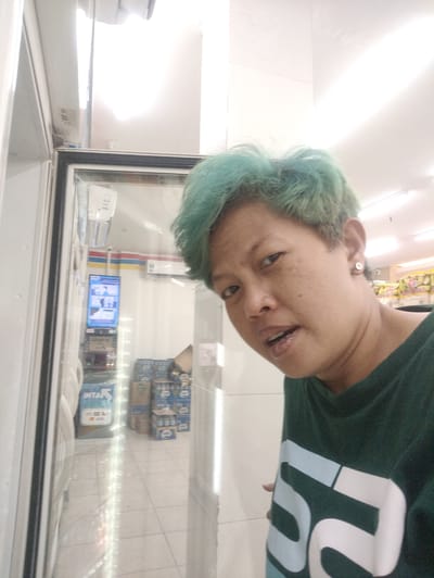 This is a selfie of a person with short, teal-colored hair. The person is wearing a dark green t-shirt with a large white and light blue graphic design on it. They have a stud earring in their right ear. The person is in a brightly lit indoor environment, likely a convenience store.
In the background, through a glass door, several stacks of bottled beverages in blue and brown packaging are visible. A digital screen displaying what appears to be instructional graphics is also visible on the wall of the store. The floor is tiled in a light color.
The lighting suggests it is daytime. The overall mood appears casual and candid. There is text visible on the blue packaging of the bottled drinks, and a white label with text that is partially obscured.