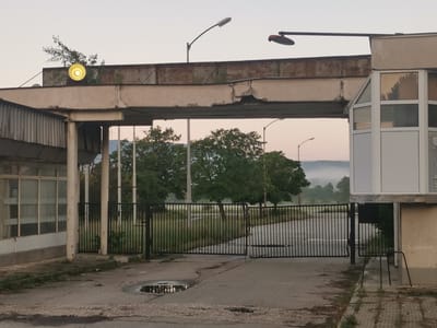 The image captures a dilapidated entrance gate and overhead structure in Zhivkovo, Bulgaria, likely depicting an abandoned industrial or administrative complex. The scene is devoid of people and human activities, conveying a sense of stillness and neglect.
In the foreground, a closed black metal bar gate blocks entry, leading to an overgrown area with several lush green trees. The ground in front of the gate is cracked asphalt or concrete, featuring a small puddle that reflects the pale sky. To the left, part of an old building with large windows is visible, while to the right stands a small, white-framed guard booth or office, also appearing weathered.
The most striking feature is the large concrete overhead structure spanning the entrance. It shows significant signs of decay, with crumbling concrete, exposed rough surfaces, and rust on its upper metal sections. A small tree or bush is growing out of the top of this structure, further emphasizing its disrepair. Two streetlights are mounted on the structure; one on the left emits a soft yellow glow, while another on the right is dark. Another streetlight is visible further back over the gate.
In the distant background, rolling hills or mountains are shrouded in a soft mist, suggesting either an early morning fog or low clouds. The sky above is clear, casting a gentle, ambient light over the entire scene. The overall impression is one of abandonment and the quiet passage of time. No discernible text is visible.