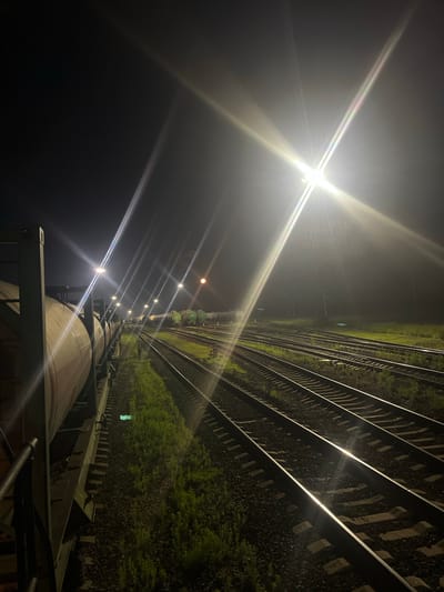 An outdoor, nighttime, long-shot photograph shows a railyard with multiple parallel train tracks. In the foreground on the left, the side of a train car, possibly a tanker car, is visible, running parallel to the tracks. The train car is light in color. The train tracks extend into the background, where several more train cars are visible, some of them green in color. The scene is illuminated by bright artificial lights, causing significant lens flare, particularly in the upper right quadrant of the image, and creating strong highlights on the train tracks. Tall light poles are positioned along the railyard. The ground beside the tracks is covered in ballast and patches of green grass. The sky is dark and black, indicating nighttime. The location appears to be a railyard or industrial area with active train lines. The scene is devoid of any human presence. The overall atmosphere is industrial and somewhat stark due to the nighttime lighting conditions.