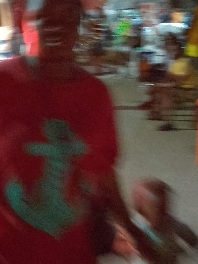 The image depicts a heavily blurred scene within what appears to be an indoor market or shop in Orie Uratta, Nigeria. An adult individual is prominently featured in the foreground, wearing a red short-sleeved shirt with a light blue or teal anchor emblem on the chest. The adult's facial features are indistinguishable due to motion blur. To the adult's right, a child figure is partially visible, also obscured by blur, positioned as if walking alongside the adult and potentially holding their hand. The child is dressed in a light-colored top. The background contains multiple indistinct figures and blurred structures consistent with market stalls or shelves stocked with various items. The ground surface is light in color. The image exhibits significant motion blur, suggesting movement, and indicates low light conditions.