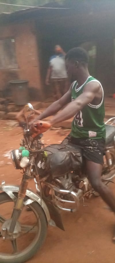 An African man in a green jersey with the number 24 is sitting on a motorcycle on a dirt road. The motorcycle has a black bag on the gas tank. In the background, there is a person standing in front of a house with a bucket. The image is somewhat blurred, suggesting movement or a low-quality camera. The ground is reddish-brown dirt, typical of rural areas.