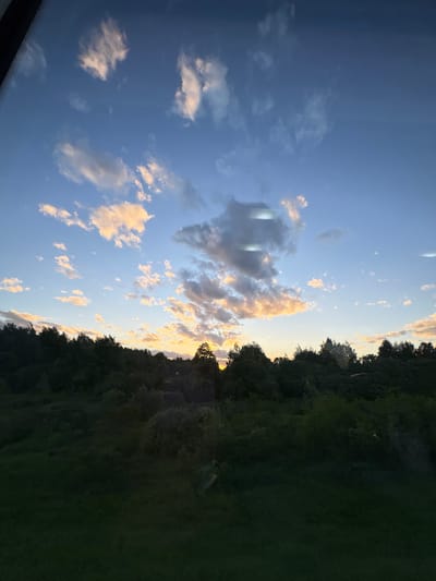 The image shows a view of the sky at Мажэйкаўскі сельскі Савет, Belarus. The sky is predominantly blue with scattered white and golden-edged clouds, suggesting either sunrise or sunset. A line of trees, likely a forest or a thick line of shrubbery, is on the right. The edge of what appears to be a dark structure or object is visible in the lower-left corner. The scene appears to be taken during a calm and possibly rural environment.