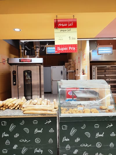 The image depicts the interior of a bakery or a section of a supermarket dedicated to baked goods. In the foreground, a display case filled with various pastries and bread items is visible. Behind the display case, a counter is laden with numerous baguettes, some wrapped in plastic. To the left, a large, stainless steel industrial oven with the brand "Forni Viorini" is prominent. The background shows more bakery equipment, including refrigerators and shelves stacked with baskets.
The overall setting is brightly lit, suggesting it is daytime. The walls are painted in warm colors, with yellow on the ceiling and a checkered pattern on the back wall. Hanging signs indicate prices and product names. One sign, in red, displays Arabic text, likely "أعز سوم" (A'az Soum), and another prominent red sign with white text reads "Super Prix". Below the counter, a dark green chalkboard-style panel is decorated with white line drawings of bread, pastries, and other baked goods, alongside text in English and Arabic, including "Pain," "Bread," "Pastry," and "مرطبات" (Murtabat - meaning pastries or sweets).
The image appears to be a candid shot of a retail environment focused on the sale of baked goods. There are no people visible in the image. The abundance of fresh bread and pastries suggests a bustling business. The lighting and the variety of products create a welcoming and appetizing atmosphere. The visible text provides information about prices, product types, and promotional offers.