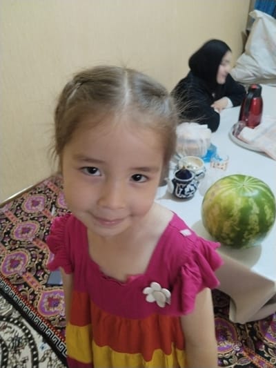 The image depicts a child in the foreground wearing a colorful dress. The child has brown hair styled in two braids and is looking at the camera. In the background, a person wearing a black hijab is seated at a table. On the table, there are various objects, including a red thermos, a watermelon, and some small containers. The environment appears to be an indoor setting with a wall in the background. The flooring is covered with a patterned rug. Based on the provided information, this scene is located in Jizzakh, Uzbekistan.