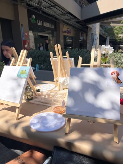 A craft setup is visible with two small easels holding canvases primed for painting. One canvas features a dog design, and the other a frog. Jars containing paint, palettes, and white cloths are also present. In the background, shops and people are seen, suggesting an outdoor setting. A person's head is near the bottom left canvas. Sunlight casts shadows across the arrangement.