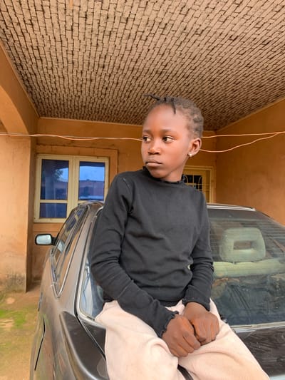 The image shows a young girl sitting on the back of a car. She is wearing a black long-sleeved shirt and light-colored pants. Her hands are clasped in her lap, and she is looking to her left with a thoughtful expression. The car appears to be parked outdoors, with a brick wall and a building visible in the background. The building has a window with blue panes. The overall lighting suggests it could be daytime, with no clear indicators of time of day or weather. The image is taken from a slightly elevated, angled perspective. No visible text is present.