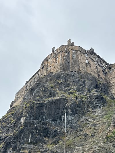 This is a low-angle, eye-level, full shot of Edinburgh Castle, situated on Castle Rock in the City of Edinburgh, United Kingdom. The castle is constructed from dark, rough-hewn stone and features numerous windows and chimneys. It is perched atop a steep, rocky cliff face that dominates the lower half of the frame. The cliff is uneven and textured, with sparse vegetation growing in its crevices. The sky above is overcast and a uniform pale gray. A single, tall, white lamppost stands on the rocky slope in the lower right quadrant of the image, providing a sense of scale. No people are visible in the frame.