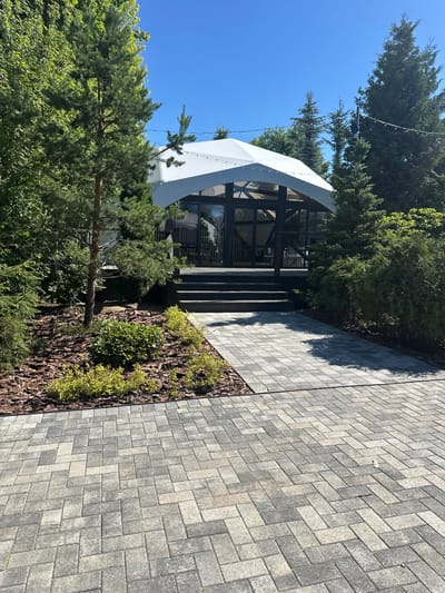 In Zakharovo, Russia, a paved walkway leads to a modern gazebo. The gazebo has a white tent-like roof and dark wooden framing with glass walls, revealing tables and chairs inside. Surrounding the gazebo are lush green trees and bushes, with mulch covering the ground in the garden bed. The walkway is made of interlocking gray pavers, creating a geometric pattern. The sky above is clear and blue, indicating a sunny day. String lights are visible along the top of the gazebo and in the trees.