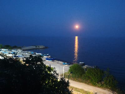 The image depicts a twilight scene at the Black Sea coast near Durankulak, Bulgaria. A full moon hangs in the deep blue sky, its reflection shimmering on the calm water. The shoreline is dotted with several small boats, mostly white and blue, resting on a sandy and rocky beach. A few of the boats are partially submerged in the water. In the foreground, lush green foliage and trees partially obscure a dirt path that curves along the coastline. A lamppost stands illuminated by the moonlight, casting a faint glow on the path. The overall atmosphere is serene and tranquil.