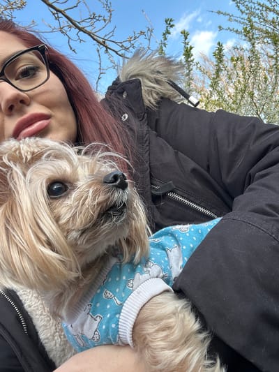 A selfie shot from a low angle depicts a person holding a small dog. The person has fair skin, auburn-colored hair, and is wearing black glasses. Their lips are visible. The dog is fluffy with tan fur and wearing a light blue shirt with a white pattern. Both are positioned close to a black jacket with a fur-lined hood. The background shows a blue sky and tree branches with budding leaves. There is no indication of the location being Gromshin, Bulgaria, or any other city, present in the image.